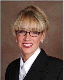 A woman with blonde hair wearing glasses and black jacket.